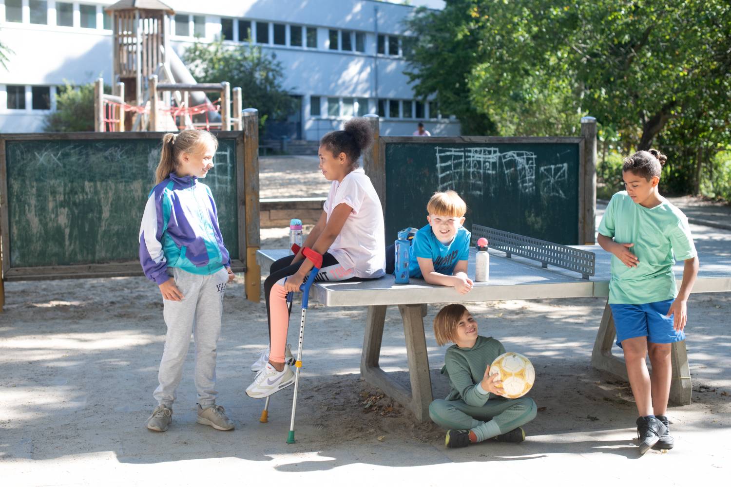 Fünf Kinder stehen an und sitzen auf einer Tischtennisplatte vor einem Gebäude mit Spielplatz und Baum