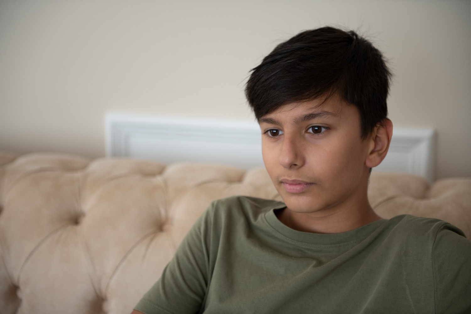Ein Junge mit dunkler Hautfarbe und Kurzhaarschnitt sitzt auf einem beigen Sofa vor einer hellen Wand und blickt nachdenklich