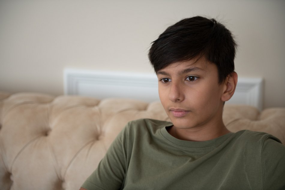 Ein Junge mit dunkler Hautfarbe und Kurzhaarschnitt sitzt auf einem beigen Sofa vor einer hellen Wand und blickt nachdenklich