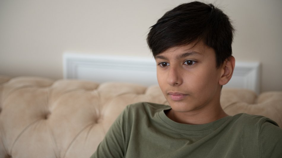 Ein Junge mit dunkler Hautfarbe und Kurzhaarschnitt sitzt auf einem beigen Sofa vor einer hellen Wand und blickt nachdenklich