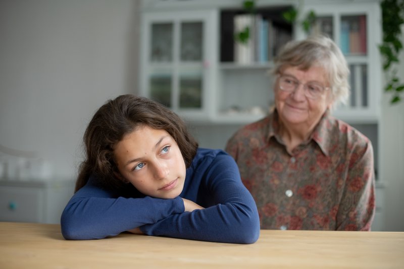 Ein junge Frau hat den Kopf auf ihre verschränkten Arme gelegt, sie ruhen auf einem Tisch. Hinter ihr sitzt ihre Großmutter und schaut sie nachdenklich an.