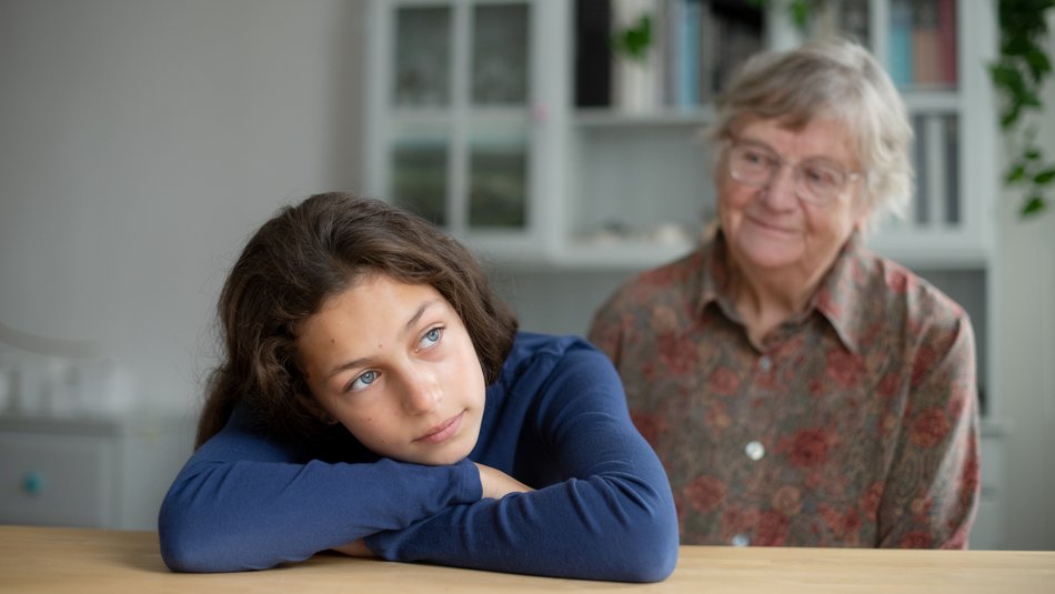 Ein junge Frau hat den Kopf auf ihre verschränkten Arme gelegt, sie ruhen auf einem Tisch. Hinter ihr sitzt ihre Großmutter und schaut sie nachdenklich an.