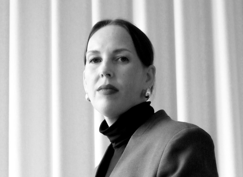 Schwarzweiß-Aufnahme von Dr. Jelena Büchner-Spielberger, die leicht von oben nach unten in die Kamera über die Schulter schaut, dunkle glatte lange hinten fxierte Haare tragend, halbrunde metallene Ohrringe und einen Blazer mit Halskragenshirt darunter