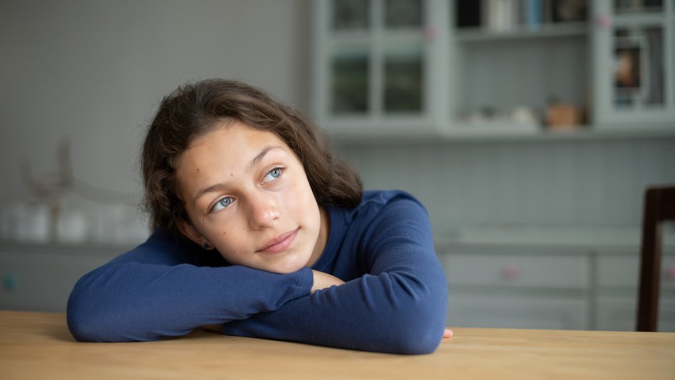 Ein Mädchen lehnt mit verschränkten Armen auf dem Esstisch und blickt nachdenklich in die Ferne