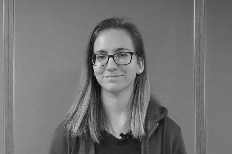 Junge Frau mit dunkler Rahmenbrille und offenen langen hellen glatten Haaren schaut lächelnd in die Kamera