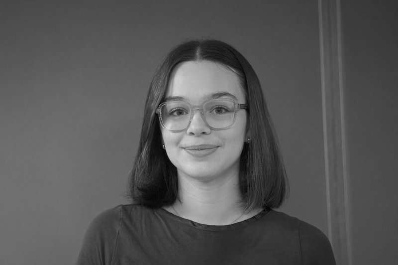 Schwarz-weiß-Foto zeigt ein junges Mädchen mit Bobschnitt, das mit dunkel gerahmter Brille in die Kamera lächelt