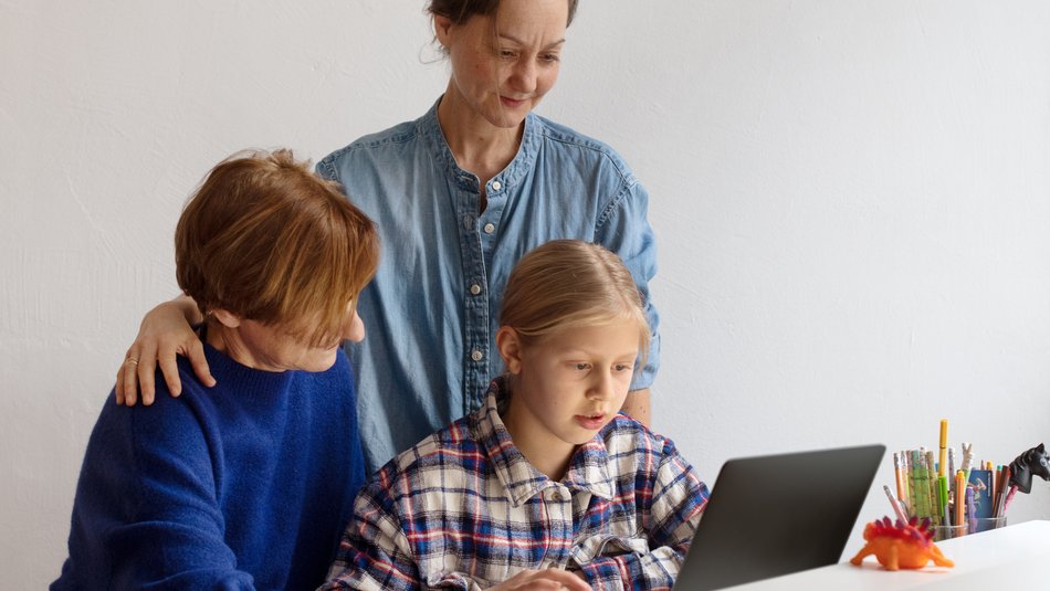 Zwei Mütter und ein Kind sitzen bzw. stehen vor einem Laptop und schauen auf den Bildschirm