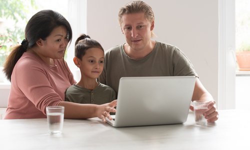 Eine Mutter und ein Vater sitzen vor einem Laptop an einem Tisch. Zwischen ihnen sitzt ihr Kind. Alle schauen auf den Bildschirm. 