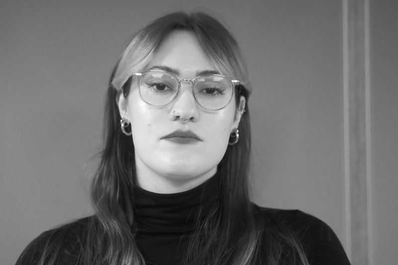 Schwarzweiß-Aufnahme von junger Frau mit gescheitelten glatten schulterlangen Haaren, Pony und Brille, die ein dunkles Rollkragenoberteil trägt und die Kamera schaut