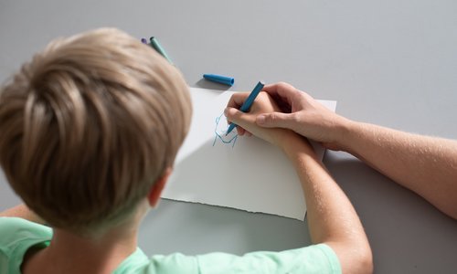 Ein Junge malt ein Bild, seine Hand wird von einer erwachsenen Hand geführt