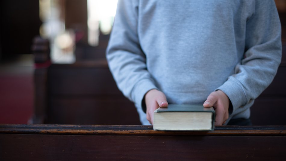 Ein Kind steht mit einer Bibel in der Hand zwischen Kirchenbänken 
