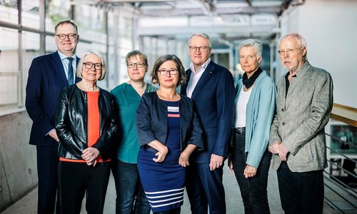 In the photo, we see the members of The Independent Federal Inquiry into Child Sexual Abuse in Germany standing side by side in a light-filled office hallway. All seven are looking at the camera.