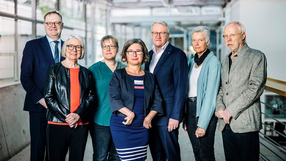 In the photo, we see the members of The Independent Federal Inquiry into Child Sexual Abuse in Germany standing side by side in a light-filled office hallway. All seven are looking at the camera.
