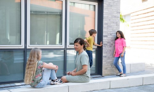 Eine Fotoaufnahme im Freien vor einer Häuserwand. Mittig sitzen ein Mädchen und eine Frau sich gegenüber, rechts daneben stehen ein Junge und ein Mädchen sich gegenüber.