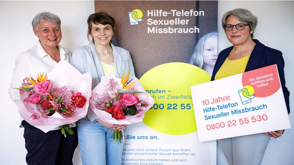 10 Jahre Hilfe-Telefon Sexueller Missbrauch Ein Gruppenfoto mit Silke Noack, Tanja von Bodelschwingh, Kerstin Claus vor einem Aufsteller "10 Jahre Hilfe-Telefon Sexueller Missbrauch".