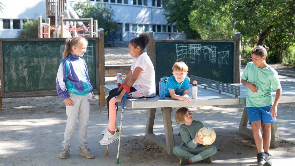 Fünf Kinder stehen an und sitzen auf einer Tischtennisplatte vor einem Gebäude mit Spielplatz und Baum