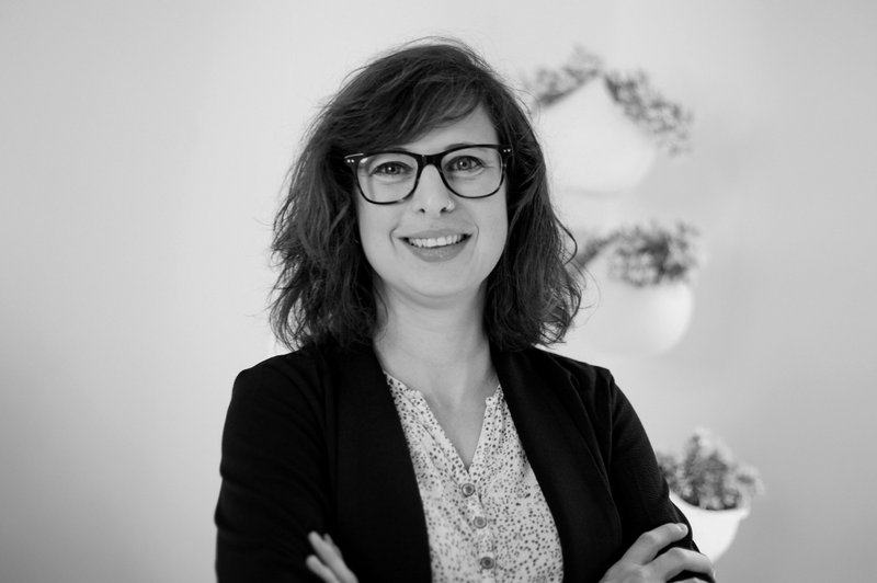 Eine Frau mit dunklem Blazer, dunkel gerahmter Brille und schulterlangen Haaren lächelt mit verschränkten Armen in die Kamera